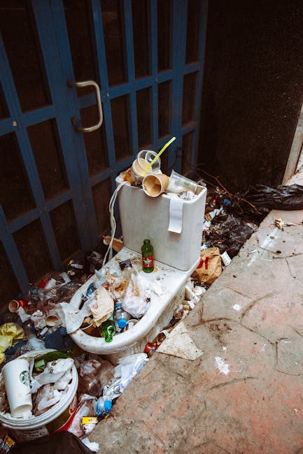 An overflowing toilet with a white ceramic fixture positioned against a dark blue metal gate, which has vertical bars and a silver handle. The toilet seat is down, and the bowl is filled with various types of litter, including plastic bottles, paper cups, and food containers. Several discarded plastic cups with straws, some tipped over, are resting on top of the toilet tank, along with a roll of toilet paper hanging over the edge. Surrounding the base of the toilet, there is a significant accumulation of rubbish, such as crumpled paper, plastic wrappers, and cans, spread across the concrete ground and nearby pavement. Additionally, a small green glass bottle is positioned upright on the toilet seat, and a black plastic bag is partially visible to the right on the darkened ground. The overall environment appears neglected and untidy, illustrating an example of improper waste disposal near a residential or commercial property, which may require professional rubbish removal services to manage effectively.