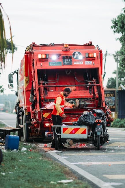 A professional waste collection worker wearing a yellow high-visibility vest and dark trousers is seen operating a large red rubbish truck on an urban street. The truck is positioned at the roadside, with its rear open for waste collection, and the worker is using tools to manage the debris inside. In the foreground, there is a black motorcycle parked close to the truck, with a black plastic bag placed on its seat. The street scene includes a paved curb, part of a green grassy area, and some scattered rubbish around the truck, indicating ongoing clearance work. The background features tall trees and a few buildings, suggesting a mixed commercial and residential environment. The lighting appears natural, with a partly cloudy sky overhead, typical of daylight hours. This scene illustrates a private waste collection service, such as Rubbish Removal Mayfair, performing on-site rubbish clearance to facilitate proper disposal outside of local authority collection, emphasizing the importance of professional private waste handling for bulky or unwanted refuse.