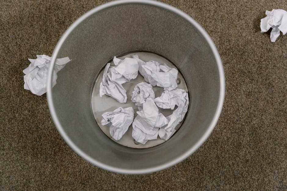 An overhead view of a cylindrical metallic rubbish bin placed on a brown textured carpeted floor. Inside the bin, several crumpled white paper balls are present, floating in a small amount of water at the bottom. A few additional crumpled paper tissues are scattered on the floor around the bin, with one piece lying slightly apart to the right. The metallic surface of the bin appears smooth and reflective, contrasting with the rougher texture of the carpet. The scene is evenly lit, emphasizing the crumpled paper’s irregular textures and the reflective quality of the bin’s outer surface, highlighting the typical setting for waste collection or disposal within an indoor environment such as an office or private premises where discreet rubbish removal practices, including on-site clearance, are relevant. This image naturally relates to private waste handling and rubbish collection services offered by Rubbish Removal Mayfair, aligning with professional waste management activities geared toward efficient and discreet disposal solutions.