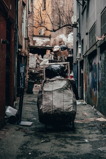 A narrow urban alleyway cluttered with a large wheeled bin covered in a weathered, beige fabric in the foreground. Behind the bin, there's a pile of mixed waste materials including cardboard boxes, plastic bags, and other debris stacked against a chain-link fence and the wall of an adjacent building. The scene is set between two buildings with graffiti on the right-side wall and a brick wall on the left, both showing signs of wear. Bare tree branches extend overhead, and the pavement is dark and slightly uneven, scattered with small debris and dirt. Visible environmental elements such as a black trash can, a fallen lid, and some discarded papers add to the cluttered appearance. The lighting suggests an overcast day, contributing to a dull and subdued atmosphere typical of an area awaiting rubbish collection or private waste disposal. This scene reflects an independent approach to rubbish removal, characteristic of private waste handling services like those offered by Rubbish Removal Mayfair, ensuring proper clearance in urban settings.