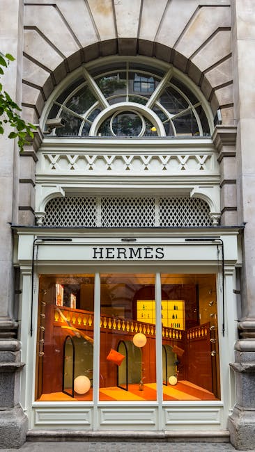 The image shows a storefront with the sign 'HERMES' on the exterior, situated beneath a decorative arched window with multiple panes, set into a stone building facade. The window features a semi-circular top with radiating muntins, framed by stone blocks arranged in a radial pattern. Below the window, there is an elaborate white architectural detail with intricate carvings and a lattice metal panel. The store's large glass display window reveals a warmly lit interior with orange-toned walls and three hanging spherical pendant lamps. Inside, various display stands exhibit handbags, including a small orange purse and other accessories, positioned on orange shelving. The storefront is clean and well-maintained, with natural daylight illuminating the facade. This visual scene reflects a high-end retail store in an urban setting, with no visible rubbish or waste management elements. Rubbish Removal Mayfair could provide services for maintaining such premises free of waste or clutter, though no waste or rubbish is depicted in the image itself.