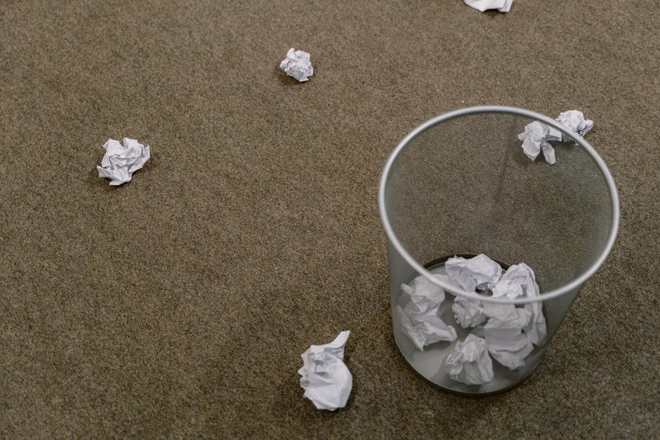 On a brown carpeted surface, several crumpled sheets of white paper are scattered around a clear cylindrical waste bin made of mesh material, which contains additional crumpled paper. The papers appear to be used and are randomly strewn, with some pieces resting on the floor near the bin. The mesh bin is positioned toward the right side of the image, and the scattering of paper creates an unorganized appearance. The scene suggests an office or workspace environment where paper waste is generated, and the image subtly reflects the need for rubbish removal or waste management services offered by Rubbish Removal Mayfair, focusing on the collection and disposal of paper waste through private or alternative collection methods.