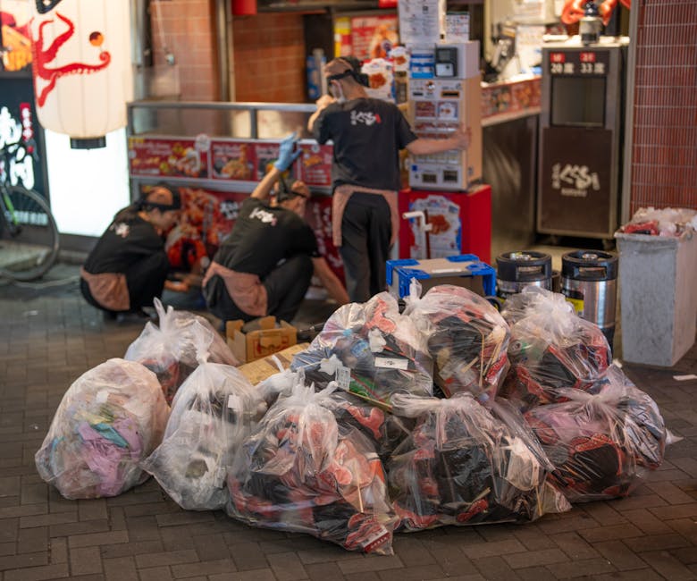 A group of three waste collection workers dressed in black uniforms with brown aprons and wearing gloves are seen working outside a commercial building, surrounded by several large transparent plastic bags filled with mixed waste, including clothing and household items. The bags are piled on the pavement near the entrance, which features a red-brick wall and a vending machine or public waste disposal bins. One worker is kneeling and appears to be handling or inspecting the waste, while another stands, possibly overseeing the collection process. The setting suggests an active on-site clearance operation, with their equipment including blue and black bins or containers, indicating an organized approach to private waste collection. The background also shows a traditional Japanese lantern hanging next to the shop storefront, which is illuminated in the evening light. This scene naturally aligns with independent rubbish removal services providing tailored waste disposal for shops and offices, as performed by Rubbish Removal Mayfair, ensuring efficient and professional rubbish handling in urban environments.