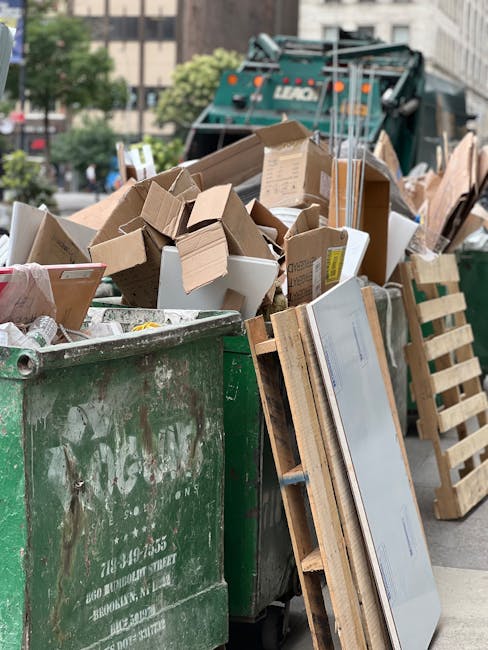 A large green waste bin is filled with an assortment of cardboard boxes, some flattened and others still in their original form, along with smaller packaging materials. The boxes vary in size and are primarily brown with corrugated textures, and some have printed labels or markings. Leaning against the bin are wooden pallets and what appears to be a flat-panel white surface, possibly a door or large appliance sheet. In the background, a waste collection vehicle with the company's branding is partially visible, parked on a city street lined with trees and multistory buildings. The scene is illuminated by natural daylight, highlighting the textures of the cardboard and wood, and indicating an active rubbish removal operation consistent with private waste collection services offered by Rubbish Removal Mayfair. The environment suggests an on-site clearance or excess rubbish disposal, supporting the context of urgent and independent rubbish clearance services for property or tenancy cleanups.