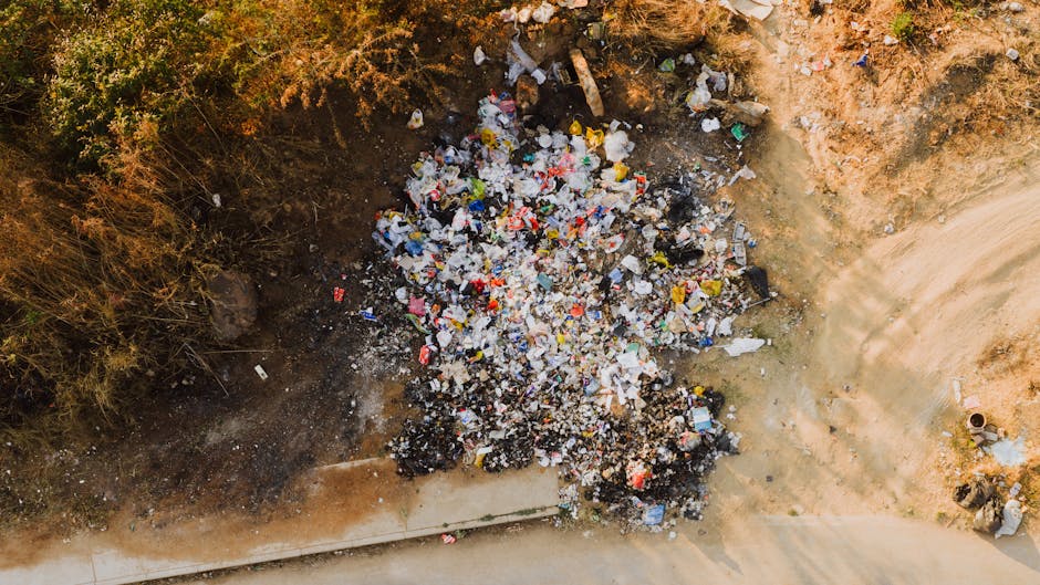 An aerial view of a large pile of mixed waste and rubbish situated on bare soil adjacent to a concrete pathway or driveway. The waste consists of various types of discarded materials, including plastic bottles, food wrappers, paper, and other miscellaneous refuse, some of which are crumpled or broken. The pile is irregularly shaped, densely packed, and appears to be recently accumulated, with some burnt or charred sections visible. Surrounding the waste on three sides are patches of dry, brown soil and sparse vegetation, including bushes and small plants. To the right, there is a cleared, dirt pathway with evident wheel tracks or footprints. The overall scene is outdoors, with natural ambient lighting illuminating the scene, highlighting the texture and color variation of the trash and earth. This image exemplifies improper waste disposal or illegal dumping, potentially addressed through private rubbish removal services such as those offered by Rubbish Removal Mayfair, in the context of such illegal dumping enforcement activities.