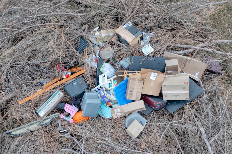 An outdoor scene showing a mound of litter and household waste scattered on dry, brown grassy ground with sparse, leafless branches in the background. The waste includes various cardboard boxes, some flattened and others intact, along with plastic containers, including a gray storage bin and a blue tray. There are black plastic trays, a small wooden crate, and a red cylindrical object partially visible among the debris. Brightly colored plastic toys and packaging materials are mixed within the waste. The materials exhibit different textures, with some smooth and shiny, others corrugated or matte. The arrangement appears haphazard, suggesting a thrown or dumped collection of rubbish possibly after an unauthorized disposal. The scene implies an instance of illegal dumping or uncontrolled waste disposal, which may involve private waste collection services like Rubbish Removal Mayfair, as indicated by the context of the webpage about enforcing proper waste management practices.
