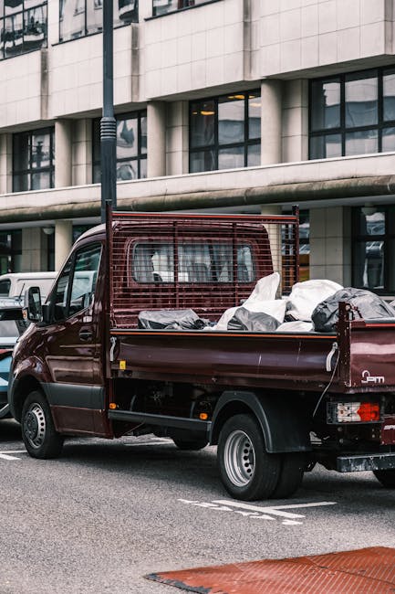 A small burgundy flatbed truck parked on an urban street in front of a modern multi-storey building with large rectangular windows. The truck’s open cargo bed contains several bags of mixed waste, including black and white plastic bags, some appearing partially filled and loosely gathered. The truck features a metal mesh guard on the cab’s rear window, colored to match the vehicle body, and its surface exhibits a slightly textured finish. The surrounding environment includes a paved road with white parking lines and a nearby sidewalk edge, with a streetlamp positioned close to the vehicle on the left side of the image. The background showcases the building's exterior with a concrete or stone facade, along with reflections visible in the windows, indicating an overcast or diffused lighting condition. This scene aligns with private waste collection services, such as those offered by Rubbish Removal Mayfair, for on-site or alternative rubbish removal tasks, especially in dense city areas where quick and efficient rubbish removal is necessary.