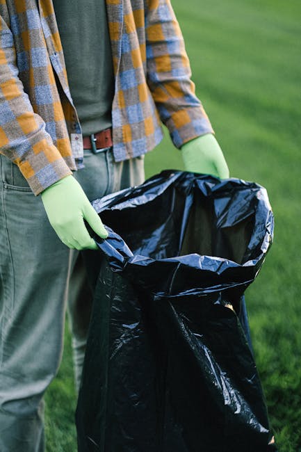 A person wearing a grey and yellow checkered jacket, grey trousers, and light green gloves is holding open a large, black plastic rubbish bag with both hands, preparing for waste disposal. The bag is situated outdoors on a grassy area, likely in a park or garden, with lush green grass visible in the background. The plastic bag appears to be sturdy and suitable for waste collection. The scene suggests an activity related to rubbish removal or garden waste disposal through private collection services. The natural lighting highlights the textures of the fabric clothing, the glossy surface of the plastic bag, and the vibrant green grass surrounding the scene, indicating daytime outdoor cleaning or rubbish clearance, possibly coordinated by a waste management company such as Rubbish Removal Mayfair. This image depicts the process of preparing waste for collection in an environment that emphasizes independent, on-site rubbish handling outside traditional waste collection infrastructure.