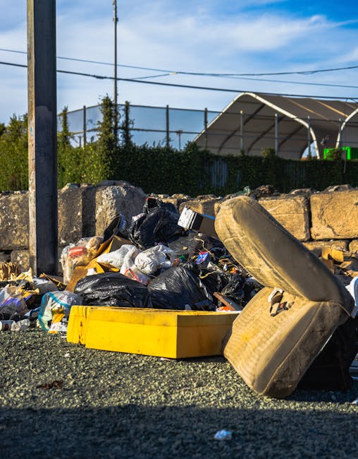 In the foreground, there is a pile of mixed rubbish including black plastic bin bags, discarded cardboard, and various small waste items, all placed on a gravel surface. Prominently visible is an old, dirty, beige car seat that appears worn and stained, positioned at an angle leaning against the rubbish pile. A bright yellow plastic container lies partially buried among the waste, with its open top facing upward. Behind the waste, a low stone wall made of irregularly shaped, weathered stones separates the rubbish area from the background. The background includes a hedge with dense green foliage, a metal fence, and a large, white, semi-circular canopy structure under a clear blue sky with a few wispy clouds. Power lines cross the scene horizontally. The overall setting suggests an outdoor area designated for waste collection or temporary rubbish storage, possibly managed by a private waste removal service such as Rubbish Removal Mayfair, supporting alternative waste handling methods outside of official municipal collection.