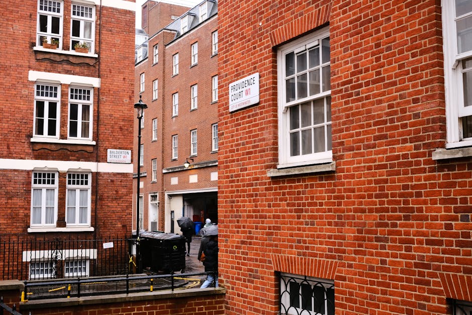 The image shows a narrow urban alleyway bordered by red brick residential buildings with large white-framed sash windows. In the foreground, on the right side, a red brick wall features a window with barred security grilles and a small flat roof above it. On the left side, a similar brick building has a row of three windows on each floor, with some flowerpots visible on the window sills. Several signs are affixed to the buildings, including street name signs indicating 'Balderton Street W1' and 'Providence Court W1.' The alleyway contains a collection of large black wheelie bins and other rubbish containers, some with lids open, positioned near a black wrought iron fence that encloses a small patio area. Two individuals, dressed in casual clothing and jackets, are visible in the background, possibly engaged in waste disposal or clearing activities. The scene is illuminated with natural daylight, with a slightly overcast sky suggesting typical urban waste management or rubbish removal activities in Mayfair, and subtly relatable to private or independent collection services provided by companies such as Rubbish Removal Mayfair.