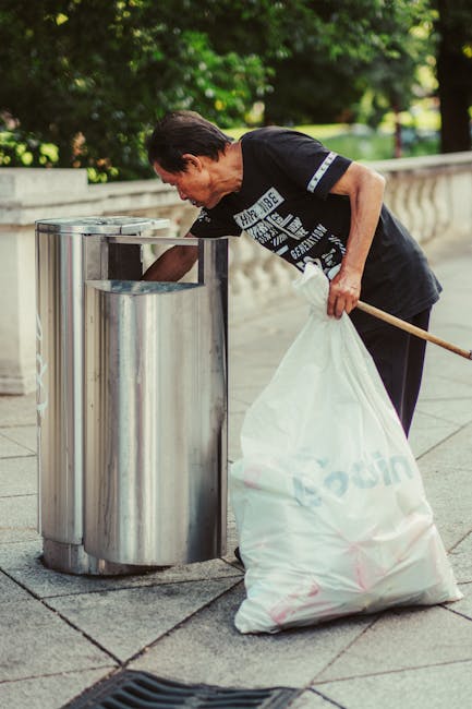 A middle-aged man with dark hair, wearing a black T-shirt with white text and dark trousers, is seen disposing of trash into a cylindrical stainless-steel public rubbish bin on a paved outdoor walkway. He is bent over slightly, holding a long wooden stick to help guide a large white plastic bag filled with rubbish into the top opening of the bin. The trash bag is partially translucent, revealing mixed waste inside, and appears to be tied at the top. The background features lush green trees and bushes, suggesting a park or urban outdoor setting, with sunlight filtering through the foliage. The scene reflects a dedicated effort at waste disposal in a public space, which aligns with the practice of independent rubbish collection and proper waste management. The stainless steel bin has a sleek, slightly reflective surface, contrasting with the rough texture of the paved surface and the natural environment behind. This image highlights responsible rubbish disposal, a component of effective rubbish removal services such as those provided by Rubbish Removal Mayfair.