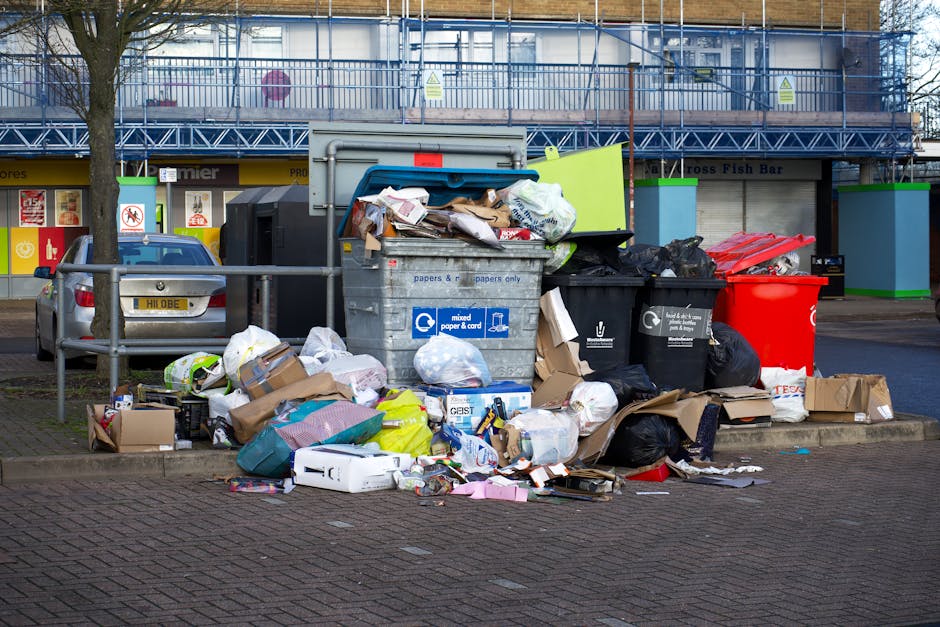A large collection of mixed waste and rubbish is piled on a paved pavement area in front of a commercial building, with several overflowing black and black-and-white recycling bins, a red bin, and a blue lid bin prominently in view. The waste includes cardboard boxes, paper, plastic packaging, and various discarded items, some crumpled or unfolded, spilling onto the surrounding area. Behind the rubbish, there is a parked silver car with a registration plate visible, and a waist-high metal railing separates the waste from the street. The background features a storefront with window displays, and a blue scaffolding or construction barrier runs along the upper level of the building, indicating ongoing work. The scene depicts an unsolicited outdoor rubbish accumulation, typical of private waste collection or refuse left for independent rubbish removal services, with natural daylight illuminating the scene clearly, emphasizing the disorderly state of the discarded materials on the pavement.