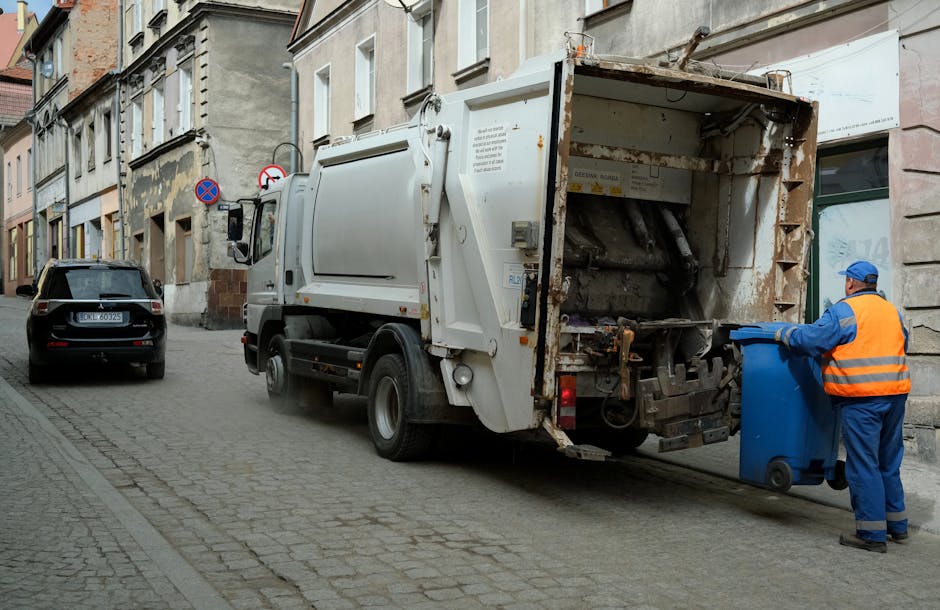 A sanitation worker dressed in a blue uniform and an orange high-visibility vest is standing on a cobblestone street, emptying a blue wheeled bin into a large, white waste collection truck. The truck has an open rear hopper with visible rust and mechanical components, indicating it is in the process of collecting rubbish. In the background, there are older, multi-story buildings with weathered facades and variously sized windows, typical of an urban residential area. A black car is parked nearby, and street signs including a no-parking sign are mounted on the buildings. The scene is captured during daytime with natural lighting, and the environment suggests a typical urban waste collection operation as part of private rubbish removal services. Rubbish Removal Mayfair routinely conducts on-site waste collection, exemplified by the activity shown in this street scene, emphasizing the direct handling and disposal of rubbish compared to public collection methods.