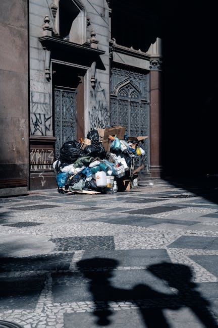 A large collection of mixed waste and rubbish is piled on a paved pavement area in front of a commercial building, with several overflowing black and black-and-white recycling bins, a red bin, and a blue lid bin prominently in view. The waste includes cardboard boxes, paper, plastic packaging, and various discarded items, some crumpled or unfolded, spilling onto the surrounding area. Behind the rubbish, there is a parked silver car with a registration plate visible, and a waist-high metal railing separates the waste from the street. The background features a storefront with window displays, and a blue scaffolding or construction barrier runs along the upper level of the building, indicating ongoing work. The scene depicts an unsolicited outdoor rubbish accumulation, typical of private waste collection or refuse left for independent rubbish removal services, with natural daylight illuminating the scene clearly, emphasizing the disorderly state of the discarded materials on the pavement.