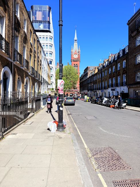 A clear urban street scene in Mayfair under bright daylight, featuring a row of Victorian-style brick terraced buildings with arched windows and iron railings along the left sidewalk. In the foreground, there is a black pole with a traffic sign attached, as well as a small pile of white rubbish bags placed on the sidewalk near the pole. The street is lined with parked scooters and cars on the right side, and a few pedestrians are visible walking along the pavement. In the background, the iconic red-brick clock tower of Westminster Palace with a tall spire and clock face stands prominently against a vivid blue sky. The scene reflects a typical city environment where additional waste collection or private rubbish disposal methods, such as those managed by Rubbish Removal Mayfair, may be employed for on-site clearance or alternative waste handling of refuse generated in this area.