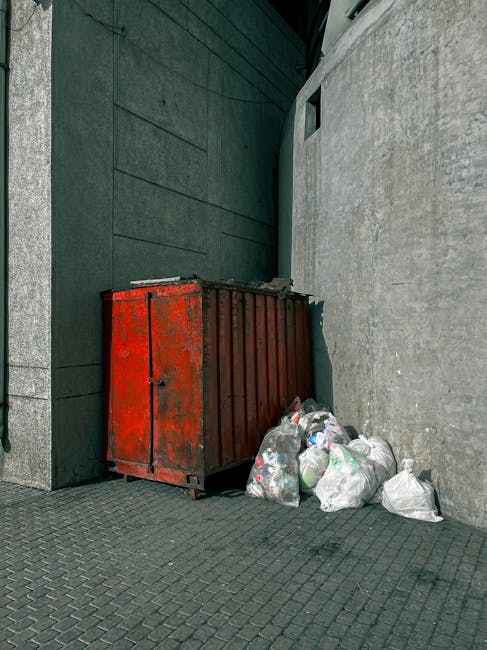 A photograph depicts a red, industrial-style metal skip with visible rust and chipped paint, positioned against a dark green wall on the left and a light grey, textured concrete wall on the right. In front of the skip, there are several loosely tied white and transparent plastic bags filled with mixed waste, including plastic bottles and packaging materials. The scene is set outdoors on a paved surface with small, dark grey interlocking tiles, and there is a narrow alleyway or corner where the walls meet. The lighting appears natural, possibly overcast, casting minimal shadows. This setup exemplifies a typical private waste removal arrangement, where an independent rubbish collection service like Rubbish Removal Mayfair could collect or handle such waste for disposal, reflecting the context of alternative waste handling outside of municipal collection systems.